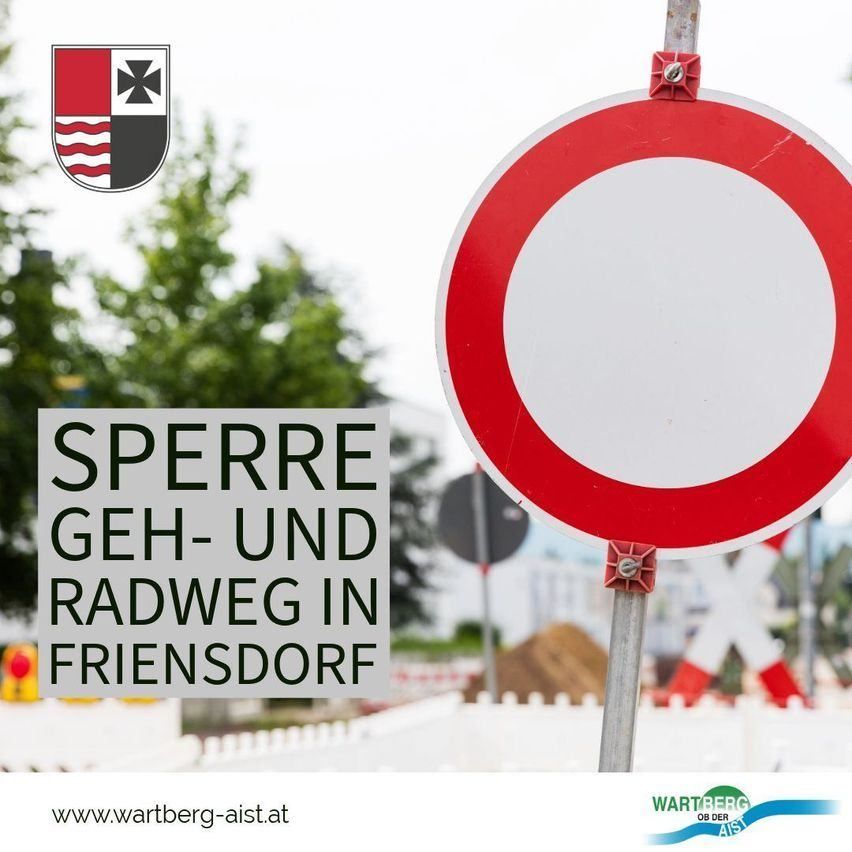 Ein rotes und weißes kreisförmiges Schild mit dem Text 'SPERRE GEH- UND RADWEG IN FRIENSDORF' ist auf einem Pfosten angebracht. Das Schild ist von einem unscharfen Hintergrund aus Bäumen und einem Logo umgeben. Das Schild befindet sich auf einer Straße mit einem weißen Zaun.