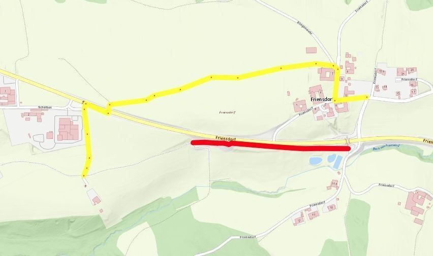 Eine Karte, die eine Straße mit einer gelben Linie und einer roten Linie zeigt, die eine andere Route anzeigt. Die Karte enthält verschiedene Gebäude und ein Gewässer.
