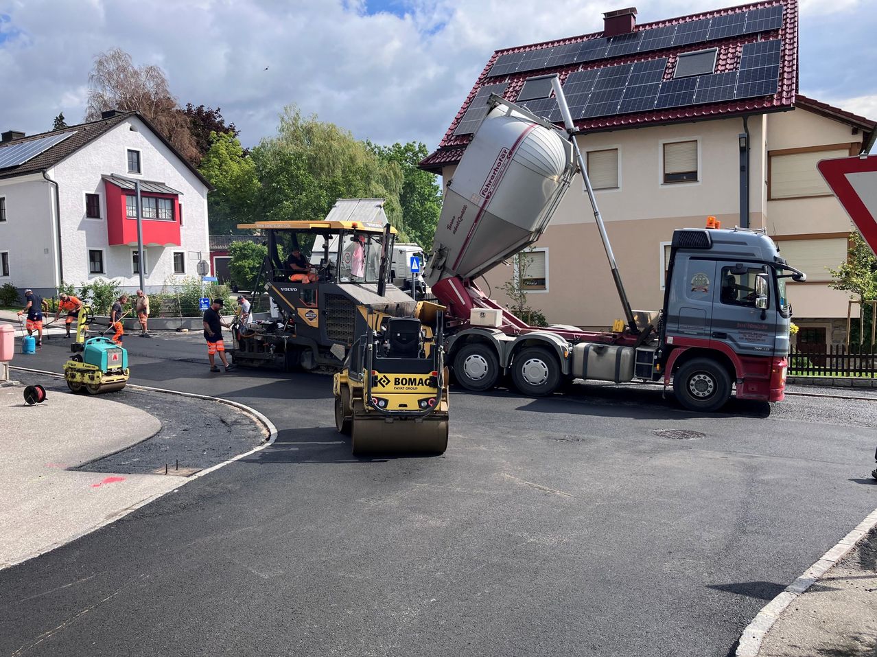 Eine Baustelle mit einem LKW, der Zement in eine Zementmischmaschine entlädt, einem Straßenwalze und Arbeitern vor einem Haus mit Solarmodulen.