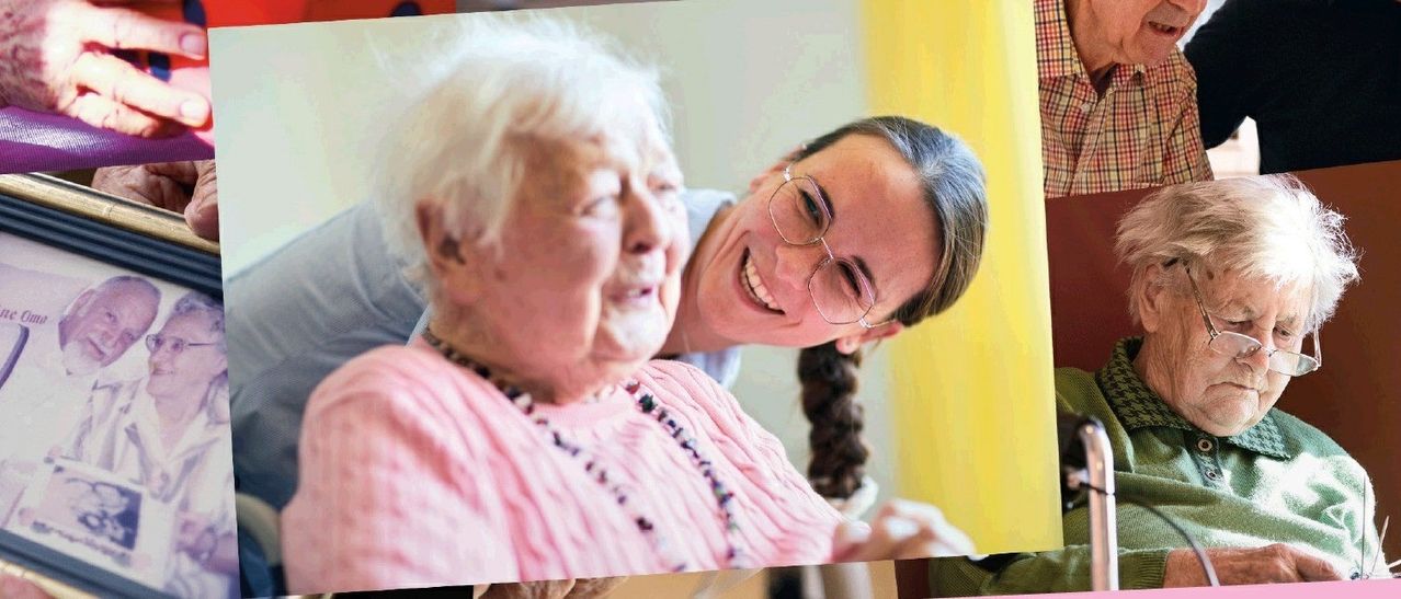 An elderly woman in a pink shirt smiles at a woman wearing glasses who is holding her shoulder.