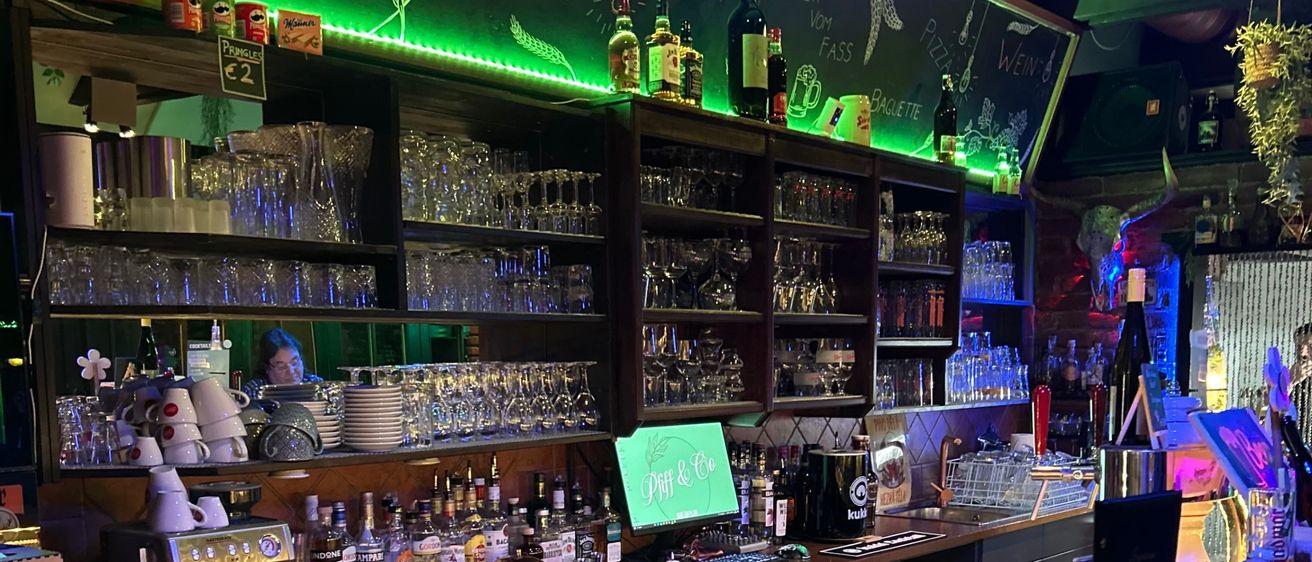 Eine Bar mit einer Kreidetafel-Menü hinter der Theke, verschiedene Flaschen und Gläser. Ein Monitor und eine Kaffeemaschine sind auf der Theke.