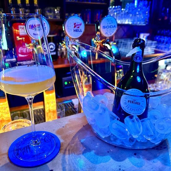 Ein Weinglas mit einem Getränk steht auf einer Bar mit einem Eiseimer und einer Flasche Perlage Wildshut Champagner.
