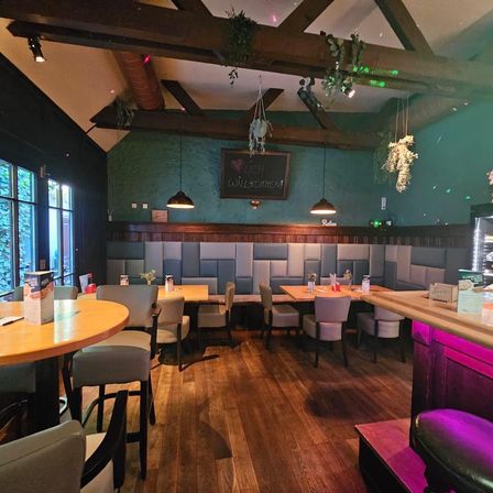 Ein Innenrestaurant mit Holzböden, grünen Wänden und Holzbänken. Es gibt hängende Lampen, eine Tafel und eine Holztheke. Die Tische sind gedeckt, und es gibt Stühle drumherum.