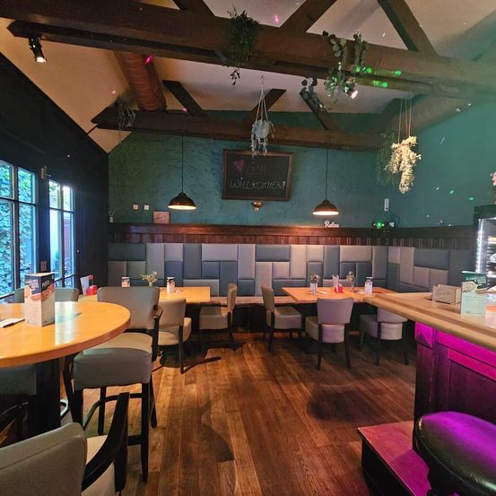 Ein Innenrestaurant mit Holzböden, grünen Wänden und Holzbänken. Es gibt hängende Lampen, eine Tafel und eine Holztheke. Die Tische sind gedeckt, und es gibt Stühle drumherum.