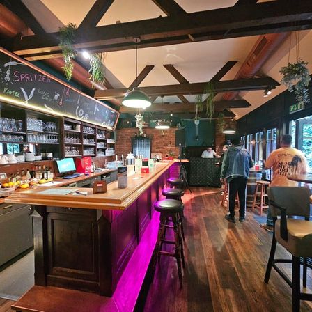 Eine Bar mit einer Holztheke, rosa LED-Lichtern und Stühlen. Eine Tafel listet Getränke auf. Ein Monitor und Flaschen stehen auf der Theke. Menschen stehen bei einem Tisch.