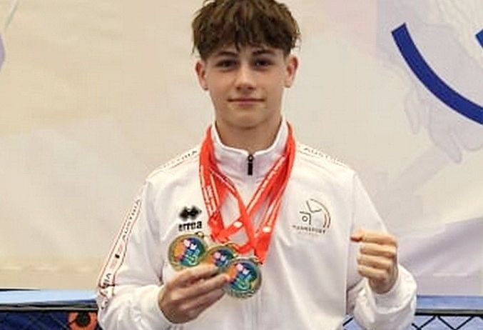 Ein junger Mann in einer weißen Jacke hält drei Medaillen, zwei goldene und eine silberne, und posiert für ein Foto.