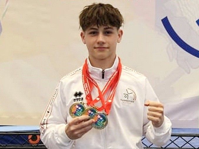 Ein junger Mann in einer weißen Jacke hält drei Medaillen, zwei goldene und eine silberne, und posiert für ein Foto.