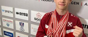 Ein junger Mann in einem roten Jacken mit mehreren Medaillen steht lächelnd vor einer Wand mit verschiedenen Logos und Bannern.