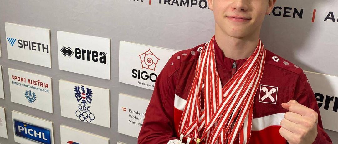 Ein junger Mann in einem roten Jacken mit mehreren Medaillen steht lächelnd vor einer Wand mit verschiedenen Logos und Bannern.