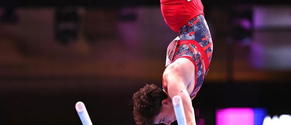 Ein Turner führt einen Handstand an den Barren aus, balanciert auf einer Hand und trägt rote und blaue Sportkleidung mit einem Logo auf der Hose.