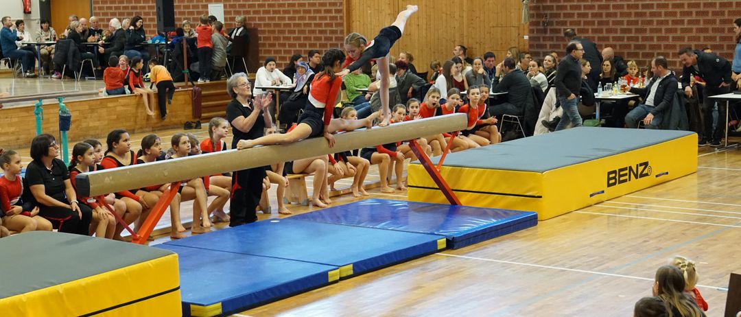Gymnasts perform on a balance beam in a gym, with spectators seated in the background. The gymnasts wear red and black uniforms. The beam is supported by blue mats.