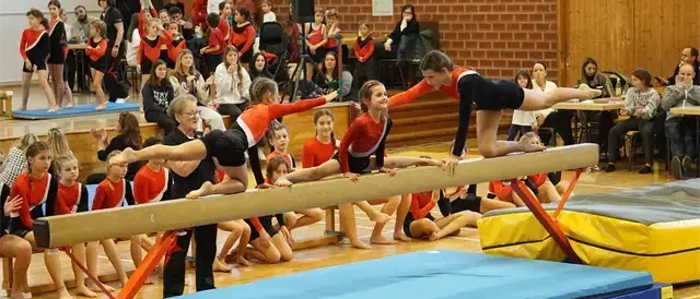 Turnerinnen in roten und schwarzen Uniformen turnen auf Sprungtischen in einer Turnhalle, während Zuschauer zuschauen. Einige Turnerinnen sind auf dem Boden, andere auf der Bank.