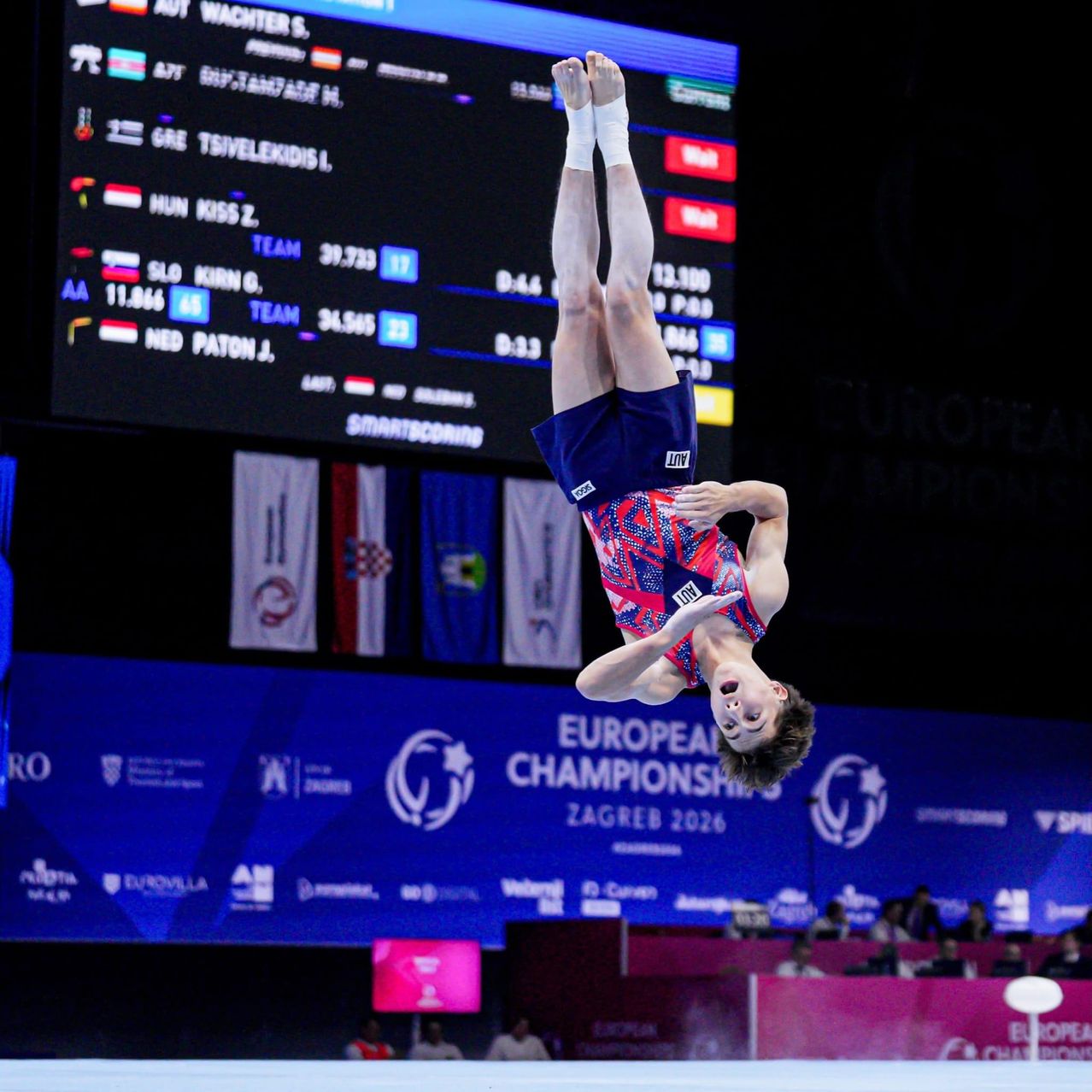 Ein Turner führt einen Handstand am Boden bei den Europameisterschaften in Zagreb durch. Die Anzeigetafel hinter ihm zeigt die Punkte verschiedener Turner.