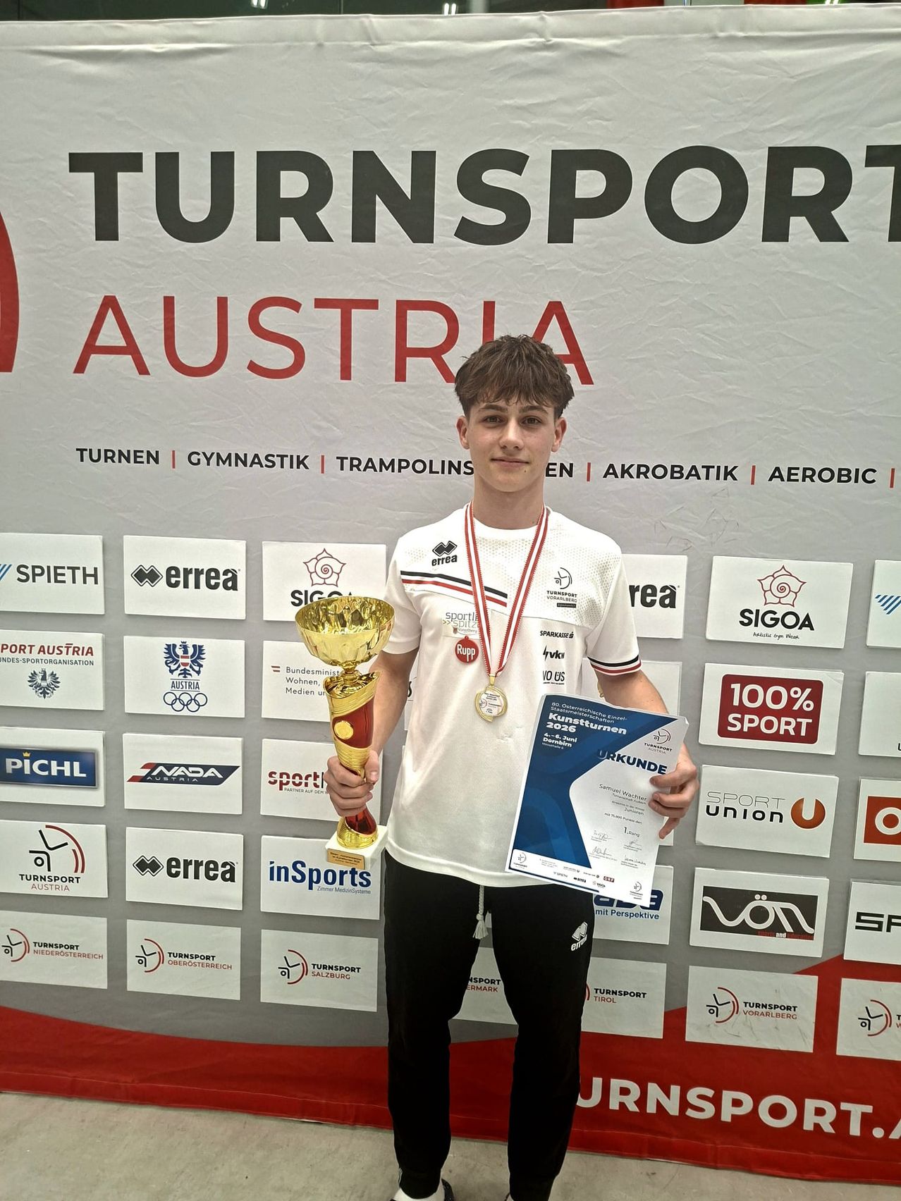 Ein junger Mann posiert mit einer Trophäe und einem Zertifikat vor einer Wand mit verschiedenen Sportlogos und dem Wort Österreich. Er trägt ein weißes Hemd und schwarze Hose.