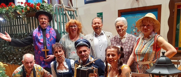 Eine Gruppe von Menschen, einschließlich Erwachsener und eines Kindes, posiert für ein Foto vor einem Gebäude mit Schildern. Einige sitzen, andere stehen, mit einem Mann, der Akkordeon spielt.