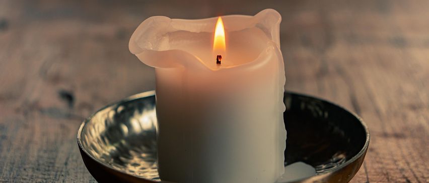 Eine brennende weiße Kerze steht in einer Metallhalterung auf einer Holzoberfläche. Die Flamme ist entzündet und das Wachs ist leicht geschmolzen.