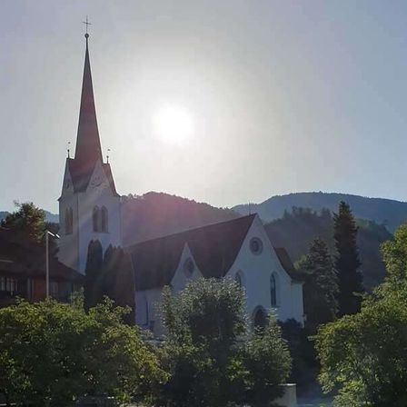 Eine Kirche mit einem Turm steht in einer üppigen grünen Gegend mit Bäumen und Bergen im Hintergrund.