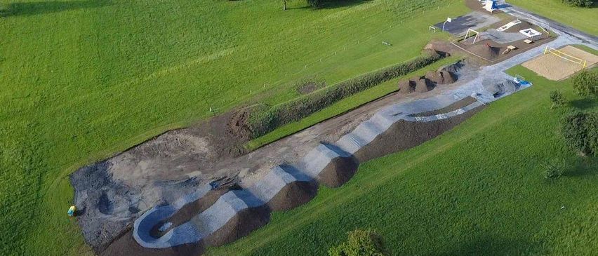 Eine Draufsicht eines grünen Feldes mit einem geschwungenen Weg aus Erde und Kies, Baufahrzeugen und einem Basketballplatz rechts.
