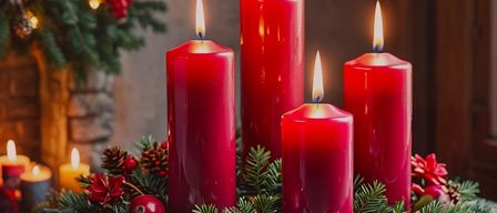 Four red candles are lit in a circular arrangement, surrounded by pinecones, red berries, and green leaves, set on a wooden table with a Christmas tree in the background.