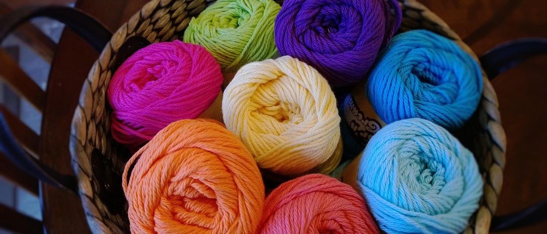 Ein Korb voller verschiedener bunter Garnrollen, darunter Rosa-, Orange-, Gelb-, Grün-, Lila- und Blautöne, ordentlich auf einer Holzoberfläche angeordnet.