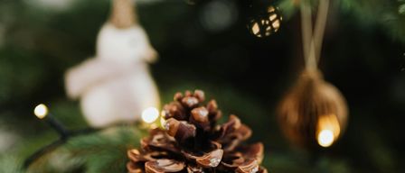 Eine Nahaufnahme eines Weihnachtsbaums mit einem Tannenzapfen im Vordergrund, hängenden Ornamenten und funkelnden Lichtern im Hintergrund.