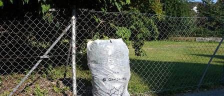 Ein Müllsack mit der Aufschrift 'Gefahren' steht neben einem Maschendrahtzaun auf dem Bürgersteig. Der Sack befindet sich am Straßenrand, dahinter ist ein Grasfeld zu sehen.