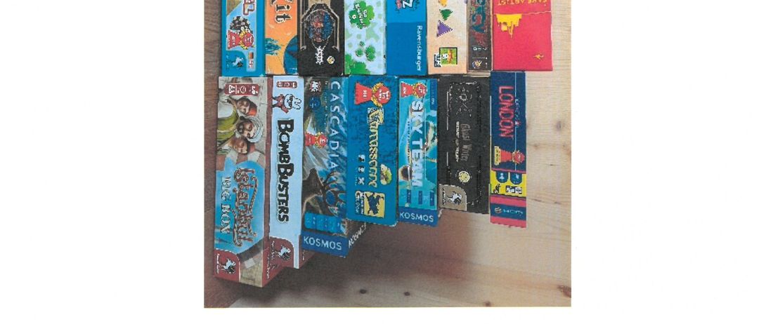 Brettspielboxen auf einer Holzoberfläche. Titel umfassen 'Flip 7', 'Dixi', 'Wortsuppen', 'Bombs' und 'Cascadia'. Ein kleines Büchlein ist ebenfalls vorhanden.