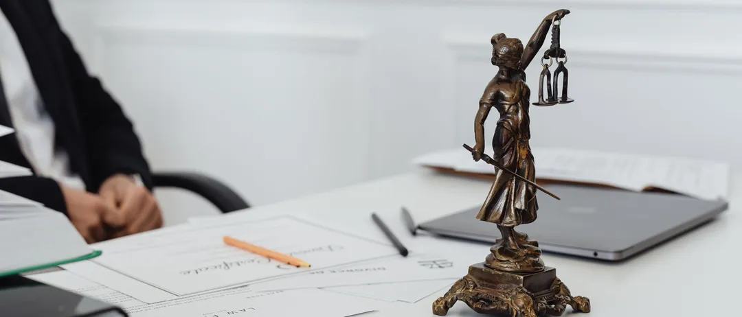 Ein Mann im Anzug sitzt an einem Schreibtisch mit einer Statue der Lady Justice, einem Laptop und verschiedenen Büroartikeln.