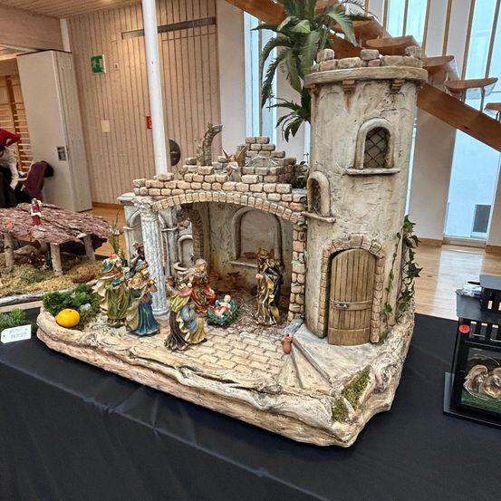 Eine detaillierte Krippe mit einem Steinbogen, Figuren von Jesus, Maria und Josef und einem Turm mit einem Bogenfenster steht auf einem Tisch mit schwarzer Tischdecke.