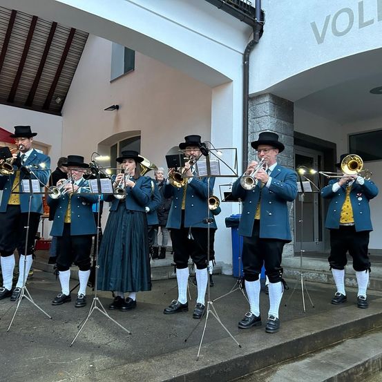 Eine Brassband in traditionellen Uniformen spielt auf den Stufen eines Gebäudes mit dem Schriftzug 'VOL'. Die Bandmitglieder halten Trompeten und Posaunen und werden von Notenständern begleitet.