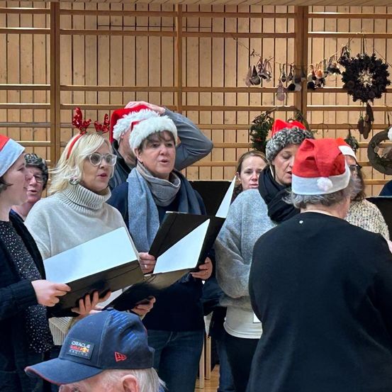 Eine Gruppe von Menschen ist versammelt, wahrscheinlich für ein Weihnachtsevent. Sie halten Bücher und tragen Weihnachtsmützen. Einige singen, während andere in ihre Bücher schauen. Die Umgebung ist drinnen mit Holzwänden und Dekorationen.