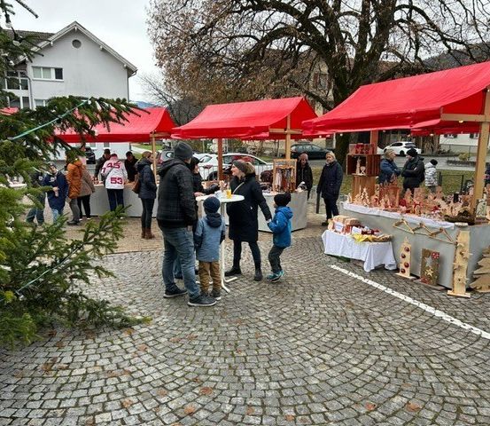 Eine Menschenmenge, einschließlich Kinder, besucht einen Weihnachtsmarkt. Drei Stände sind mit verschiedenen Artikeln unter roten Vordächern aufgestellt.