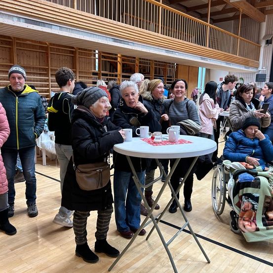 Mehrere Menschen versammeln sich um einen Tisch mit Kaffeetassen in einer Halle. Einige tragen Winterkleidung und Mützen. Eine Frau im Rollstuhl ist in der Nähe.