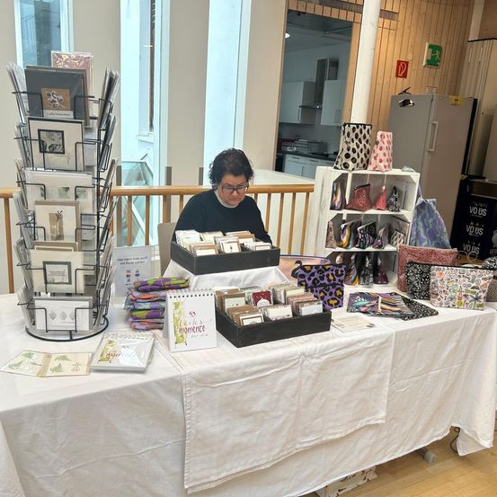 Eine Frau sitzt an einem Tisch und stellt verschiedene Bastelartikel aus, darunter Taschen, Karten und ein Notizbuch. Der Raum ist innen mit Holzwänden und großen Fenstern.