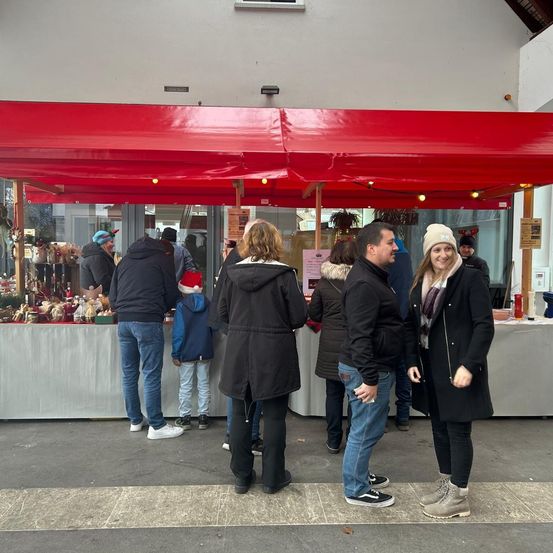 Menschen stehen um einen rot bedachten Stand, wahrscheinlich ein Markt, mit einer Ausstellung von Geschenken und Dekorationen. Eine lächelnde Frau trägt eine Mütze, und ein Mann neben ihr schaut in die Kamera. Hinter ihnen unterhält sich ein Mann mit einem Weihnachtshut mit einer Frau.