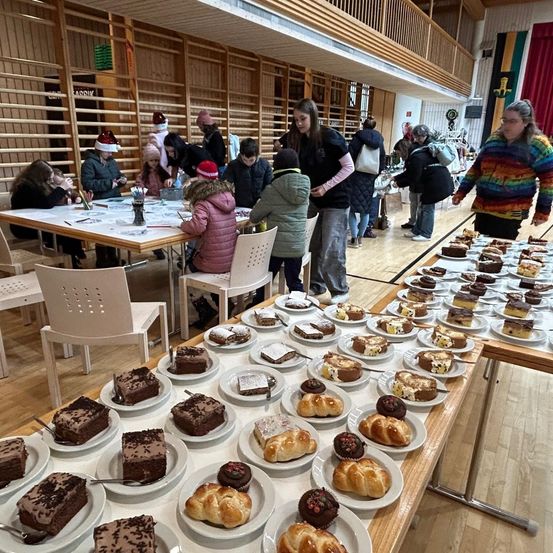 Eine festliche Zusammenkunft in Innenräumen mit Menschen in Winterkleidung. Tische sind mit einer Vielzahl von Desserts beladen, darunter Kuchen und Gebäck. Mehrere Personen sitzen und sind mit einer Aktivität beschäftigt. Die Umgebung umfasst Holzkonstruktionen und eine hohe Decke.