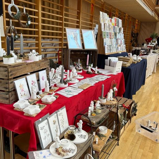 Ein Raum mit einer Ausstellung verschiedener Kunsthandwerke und Dekorationen, mit Tischen, auf denen Kerzen, Figuren und gerahmte Bilder stehen. Holzregale und Kisten verleihen dem Raum einen rustikalen Charme.