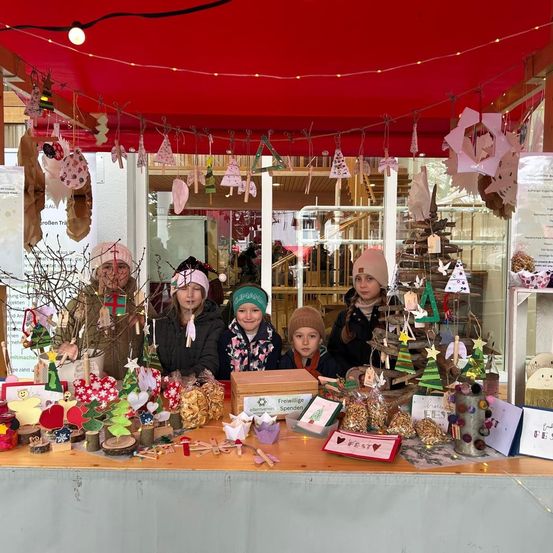 Eine Gruppe von Kindern steht hinter einem festlich dekorierten Tisch, geschmückt mit selbstgemachten Ornamenten, einem roten Baldachin und funkelnden Lichtern, was eine fröhliche Feiertagsatmosphäre schafft.