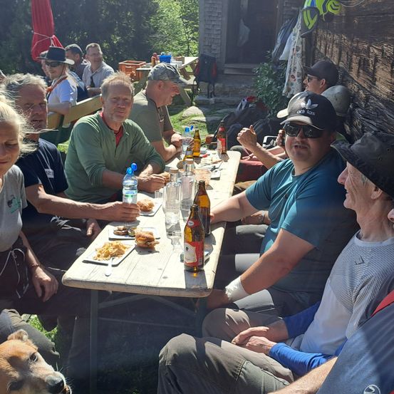Mehrere Menschen sitzen draußen um einen Tisch herum und genießen Essen und Getränke. Ein Hund ist im Vordergrund. Im Hintergrund befinden sich Bäume und ein Holzgebäude.