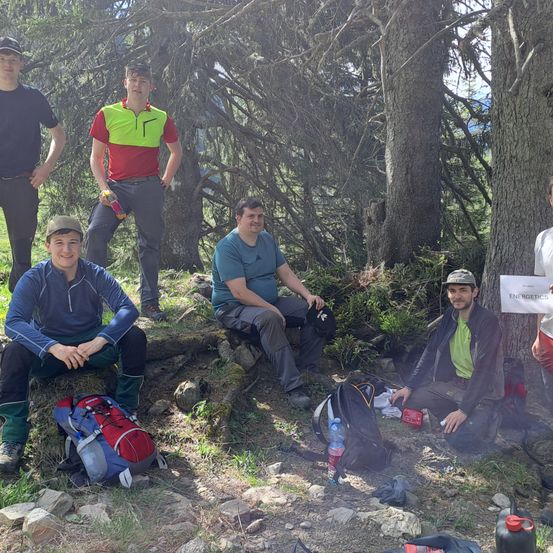 Sechs junge Männer posieren in einer Waldlichtung. Einer sitzt auf einem Baumstumpf, ein anderer hält eine Flasche. Sie tragen Rucksäcke und Mützen, einer hält ein Schild.