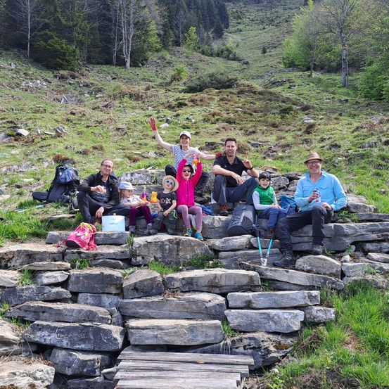 Eine Gruppe von Menschen, einschließlich Erwachsener und Kindern, sitzt auf einer Steinmauer in einem Grasbereich mit einem Berg im Hintergrund. Einige von ihnen halten Tassen.