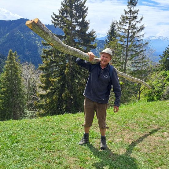 Ein Mann hält einen großen Ast über seinen Schultern auf einem Berg mit blauem Himmel darüber.