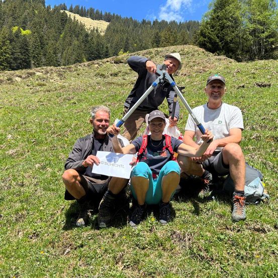 Eine Gruppe von Wanderern sitzt auf einem Grashang mit einem Schild, das G'SCHICHTL sagt. Sie alle lächeln und posieren für ein Foto.