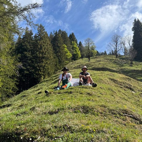 Zwei Männer sitzen auf einem grasbewachsenen Hügel und tragen Hüte und Sonnenbrillen. Einer hält eine Kettensäge und sie scheinen eine Pause einzulegen.