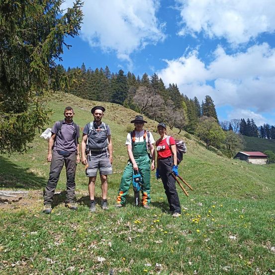 Vier Personen, die für Outdoor-Aktivitäten gekleidet sind, stehen auf einem Grashang. Sie halten Wanderstöcke und tragen Hüte, wobei Bäume und ein Haus im Hintergrund zu sehen sind.