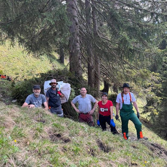 Fünf Personen posieren auf einem Hügel in einem Wald, umgeben von hohen Bäumen und Grün. Sie tragen lässige Kleidung, und eine hält einen weißen Sack.
