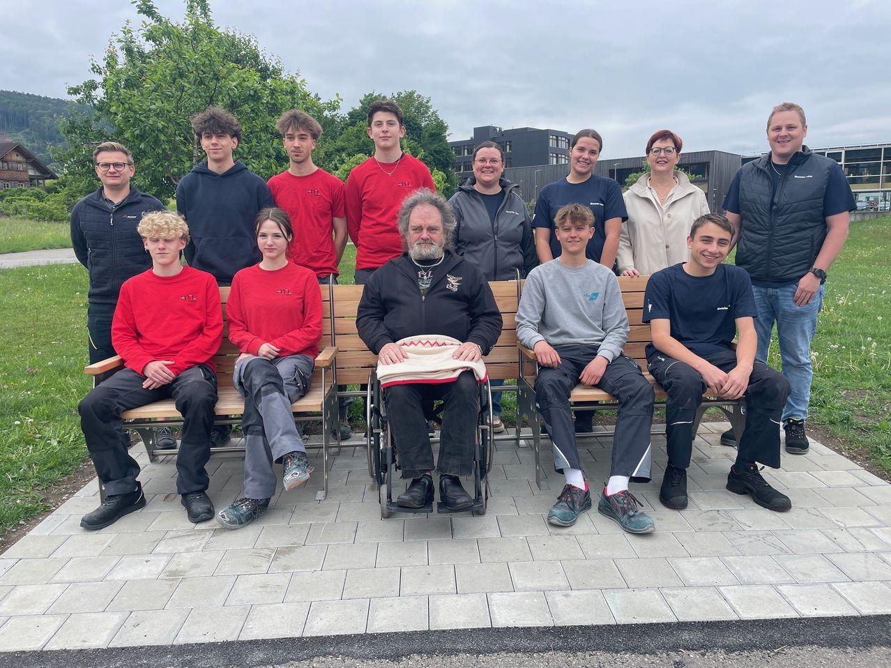 Eine Gruppe junger Menschen und Erwachsener posiert für ein Foto auf einer Bank. Bäume und Gebäude sind im Hintergrund zu sehen.