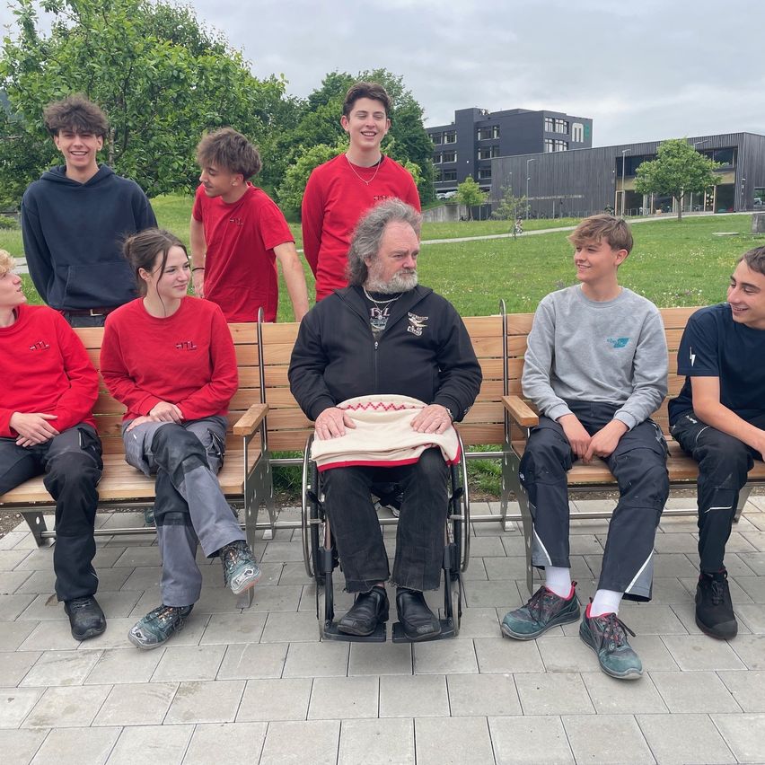 Eine Gruppe von Menschen, darunter ein Mann im Rollstuhl, sitzt auf einer Bank im Freien und lächelt für ein Foto.