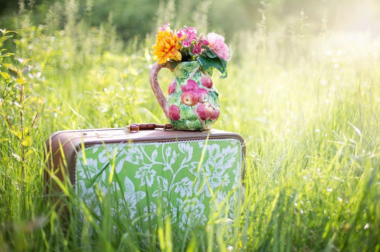 Ein Vintage-Koffer mit Blumendruck steht in einem Feld. Darauf befindet sich ein Keramikkrug, gefüllt mit rosa und gelben Blumen.