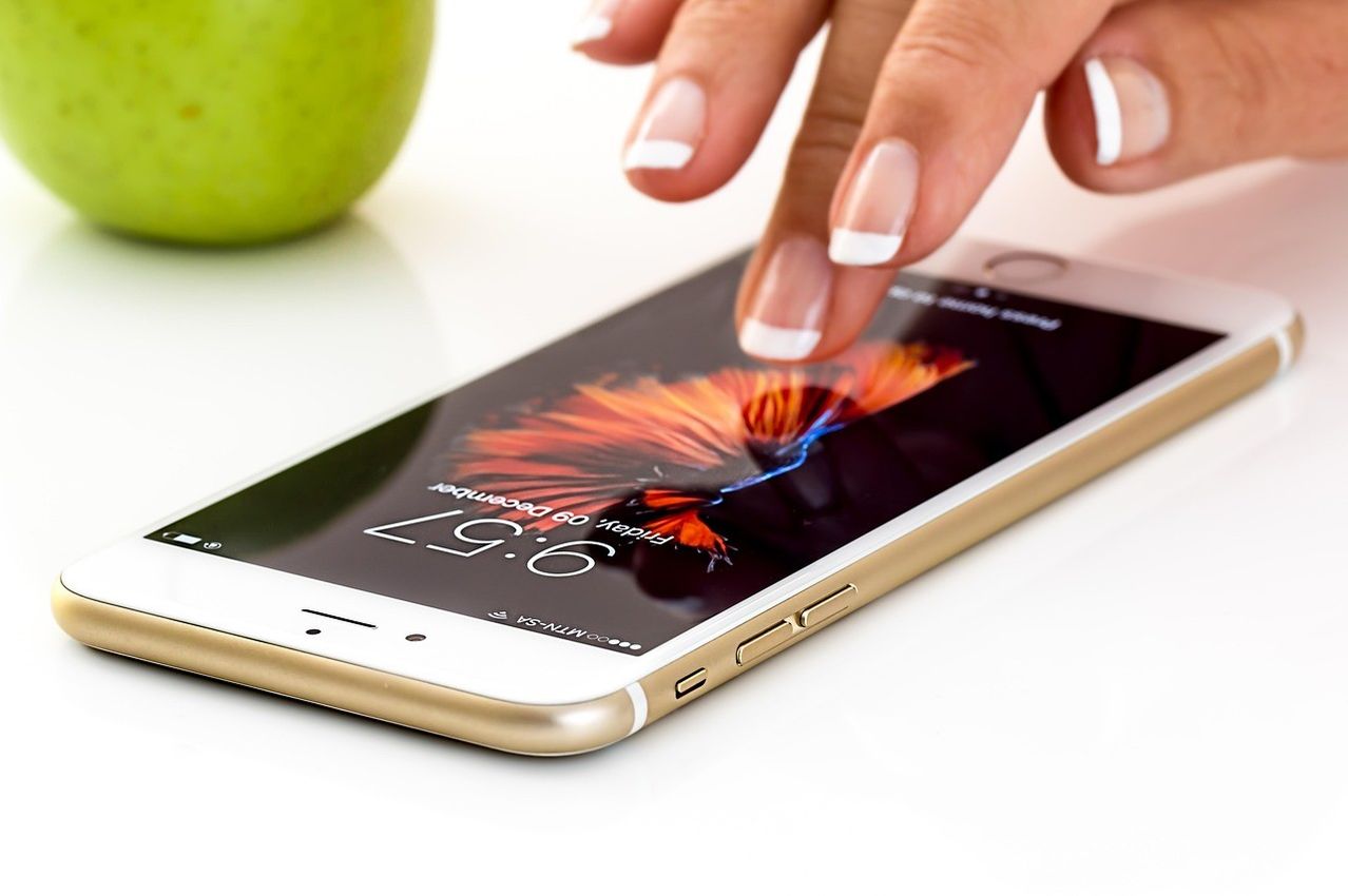 Eine Hand mit weißen Nägeln berührt ein Handy, das die Zeit 6:59 anzeigt. Hinter dem Handy steht ein grüner Apfel.